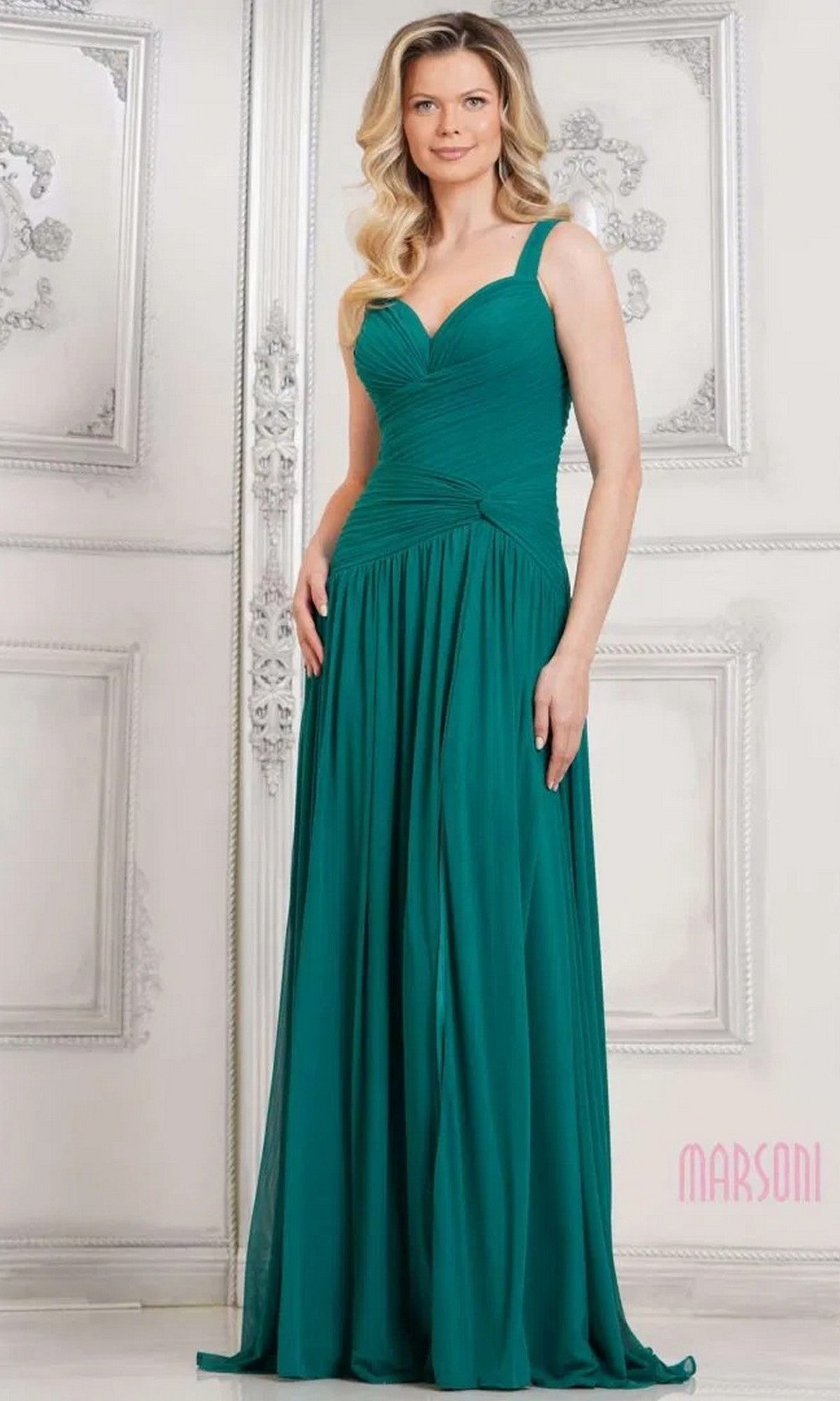 A woman in the deep green Marsoni MV1316 long formal dress with a sleeveless, ruched bodice stands before an ornately decorated white wall.
